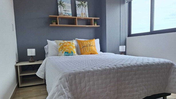 Un dormitorio con cama, dos almohadas, una mesita de noche, una lámpara, una ventana y una pared decorada con dos cuadros.