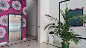 A modern hallway with a vibrant floral mural, an elevator, a potted plant, and a framed abstract painting. - Turin Airport apartment G (Leinì)