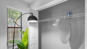 Een wasserette met een witte deur, een plant, een zwarte vloerlamp, een kledingrek met een strijkplank, en een raam met zonwering.