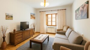 Een woonkamer met een bank, een salontafel, een televisie op een houten kast en een raam met gordijnen.