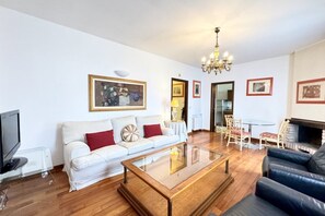 A living room with a white sofa, a glass coffee table, a black leather sofa, a chandelier, and framed pictures on the walls.