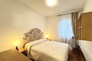 A bedroom with a bed, a nightstand with a lamp, a wooden wardrobe, and a window with curtains.