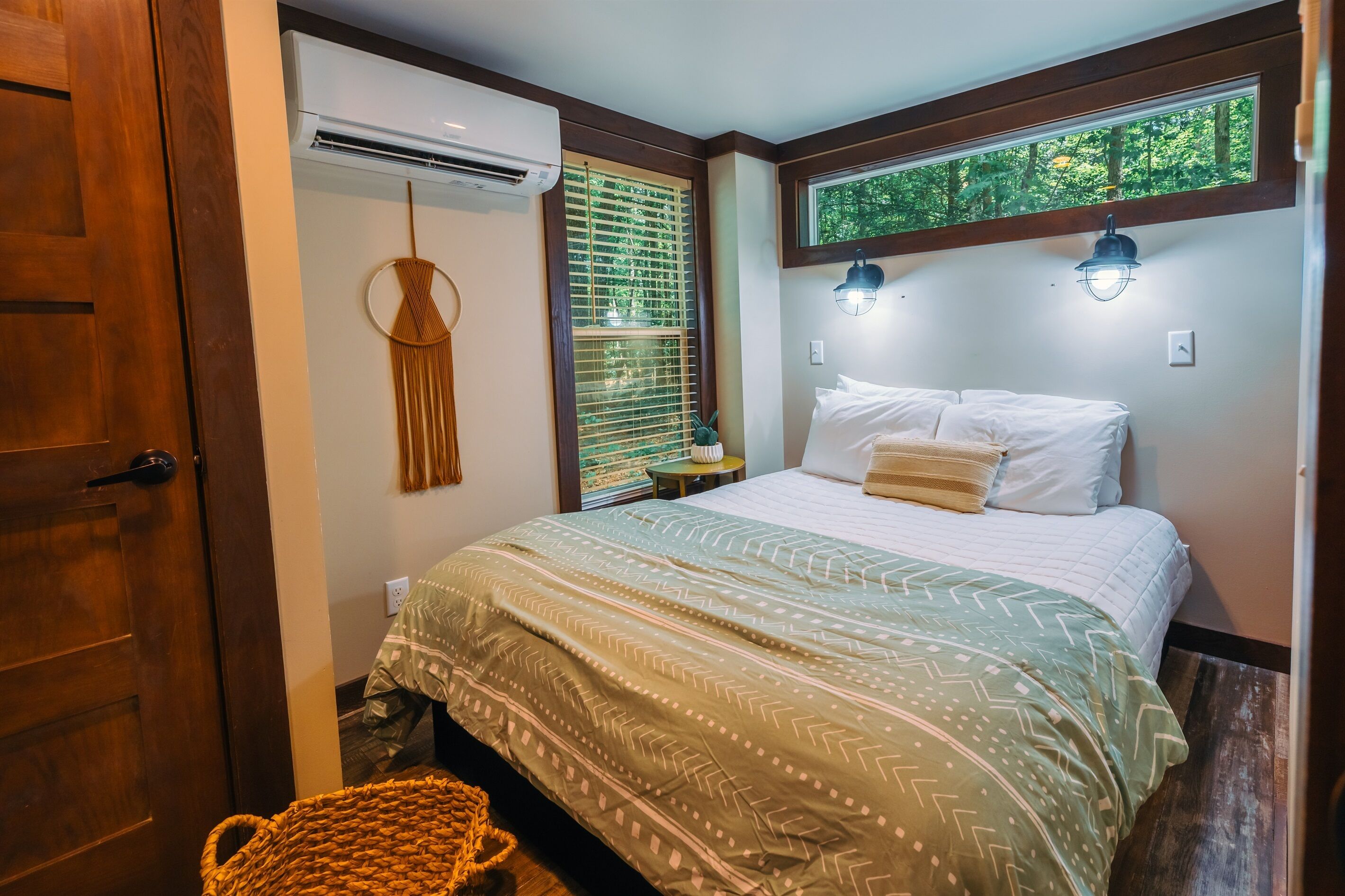 A bedroom with a bed, a hanging macrame decoration, an air conditioner, a window with blinds, and a view of trees through the window.