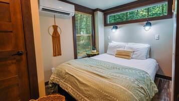 A bedroom with a bed, a hanging macrame decoration, an air conditioner, a window with blinds, and a view of trees through the window.