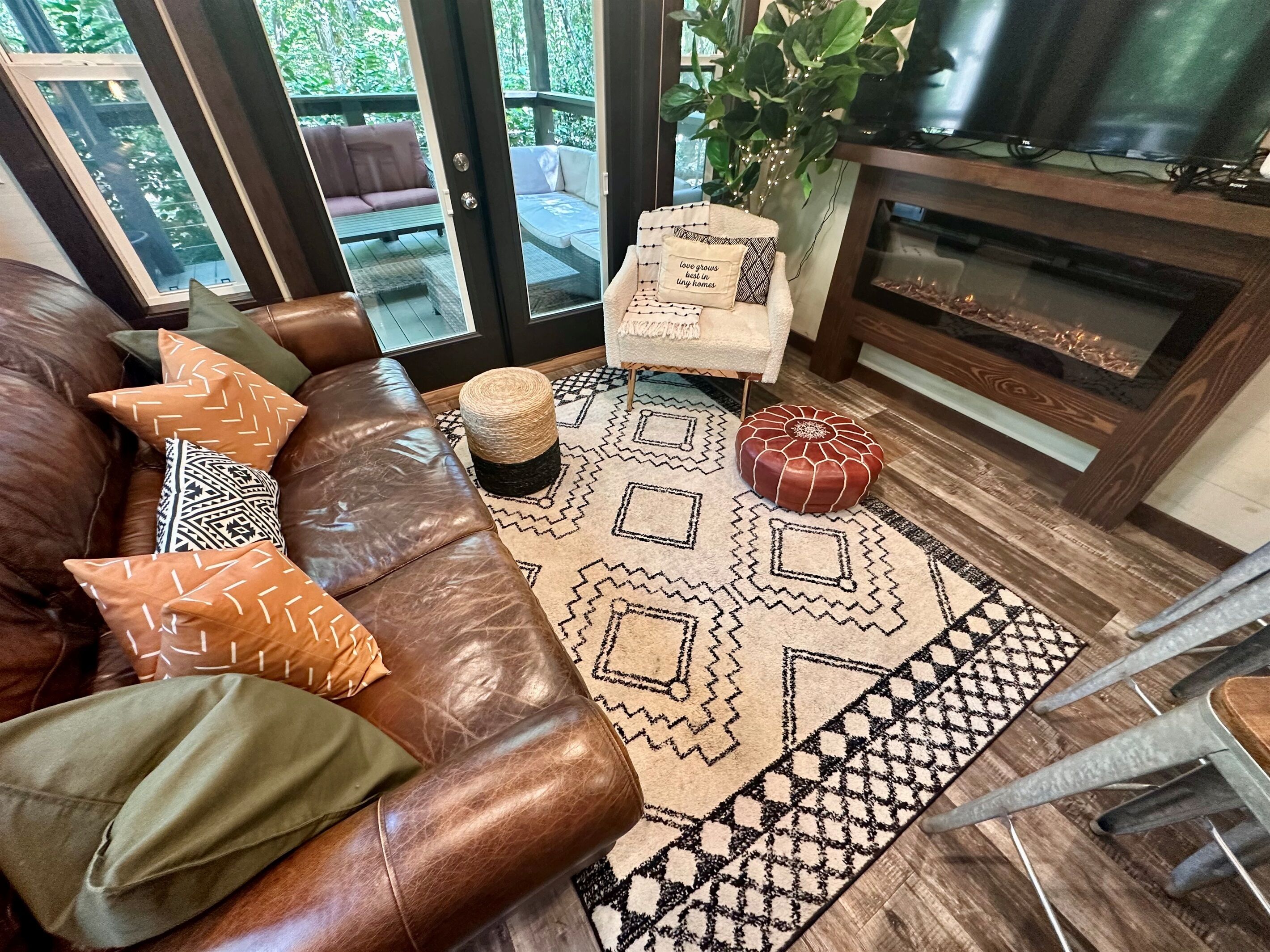 Ein gemütliches Wohnzimmer mit einem braunen Ledersofa, gemusterten Kissen, einem gewebten Hocker und einem geometrischen Teppich.