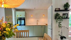 Una cocina moderna con armarios verde claro, encimera blanca y horno integrado. Hay un comedor con una mesa y sillas de madera, y una mesita blanca con una planta en maceta.