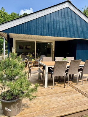 Une terrasse extĂ©rieure couverte, avec une table et des chaises, un pot de fleurs et un bĂątiment dont le revĂȘtement est bleu.