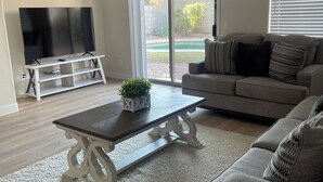 A modern living room with a flat-screen TV on a white stand, a dark coffee table with a plant, a grey sofa with patterned cushions, and a ceiling fan. - 3-bedroom house with TVs, grill, private pool and WiFi in wonderful Avondale (Avondale)