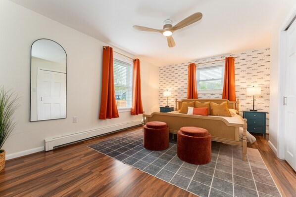 A bedroom with a bed, two ottomans, a nightstand, a ceiling fan, and a large mirror.