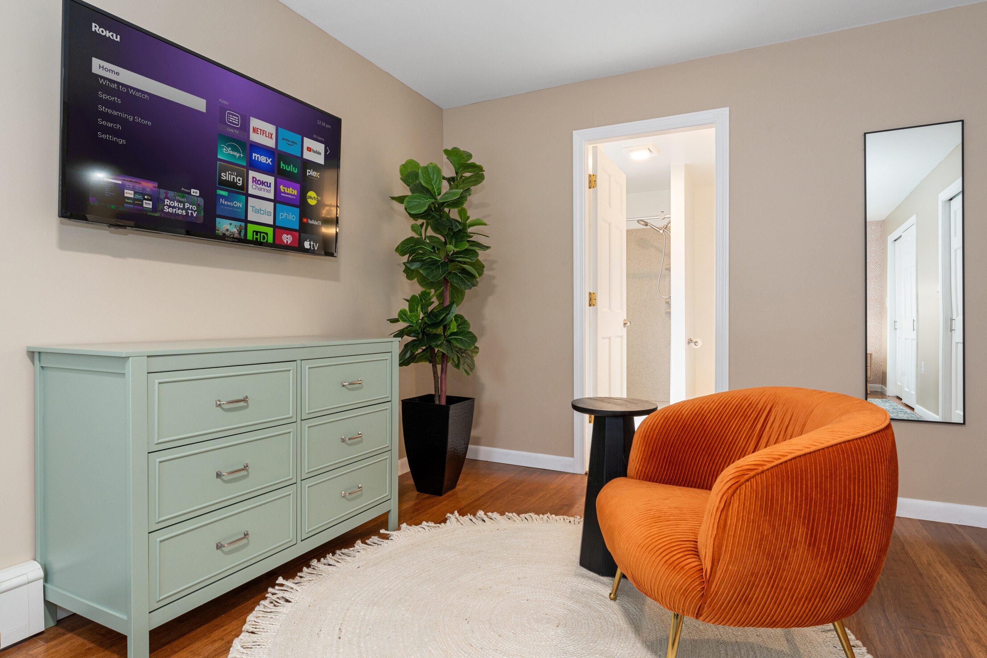 A living room with a flat-screen TV mounted on the wall displaying a streaming service interface, a green dresser, a large potted plant, an orange chair, and a white rug.