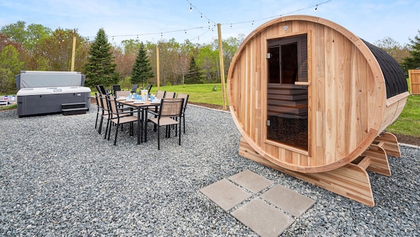 A wooden sauna with a curved exterior, a patio with a table and chairs, and a barbecue grill in the background.