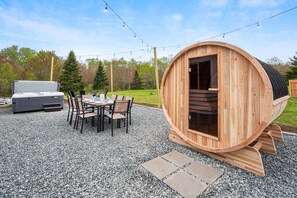 Un sauna en bois à l’extérieur incurvé, une terrasse avec une table et des chaises, et un barbecue à l’arrière-plan.