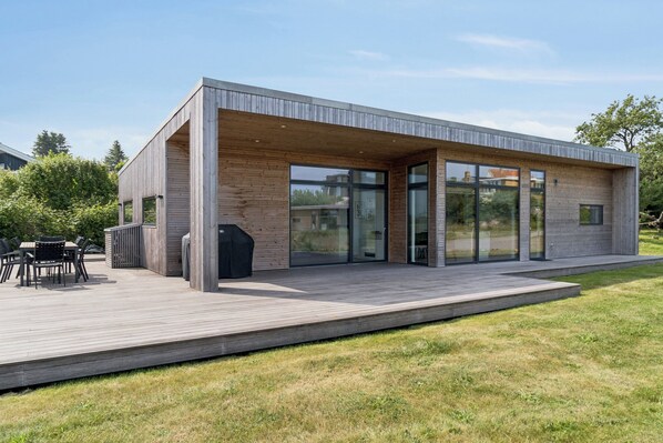 Une maison moderne dotée d’une terrasse en bois, de grandes fenêtres et d’un patio aménagé avec du mobilier d’extérieur.
