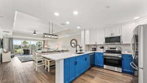 Una cocina moderna con armarios blancos y azules, una isla central, electrodomésticos de acero inoxidable y un amplio ventanal con vistas a un jardín.