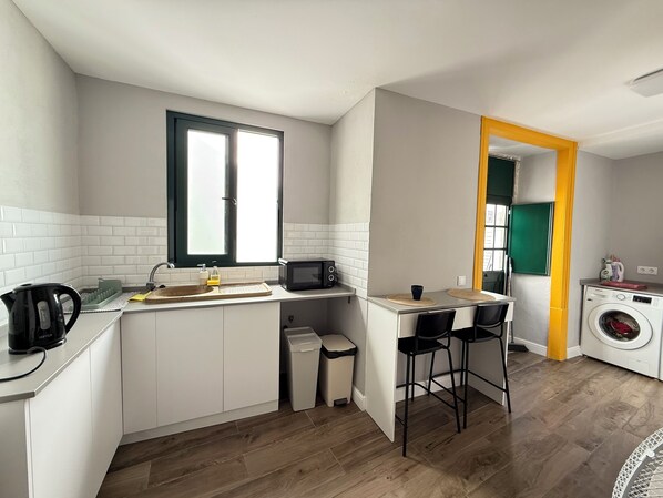 A compact kitchen with a microwave, kettle, and sink. There's a small dining area with stools and a washing machine in the corner. - Faro Coast Guest House (Faro)