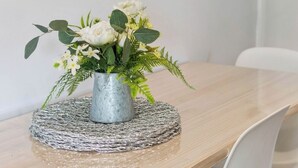 Una mesa de comedor con un florero de metal que contiene flores blancas y follaje, sobre un mantel individual texturizado.