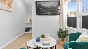 Una sala de estar moderna con un televisor de pantalla plana montado en la pared, una mesita redonda con un control remoto, un sillón verde y una planta en maceta.