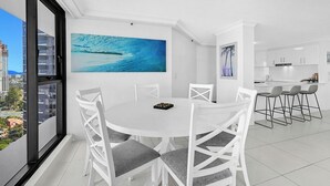 A modern dining area with a round table and white chairs, a kitchen with white cabinets, and a large window with a city view. - Stunning Ocean View 3BR near Broadbeach | Parking (Surfers Paradise)