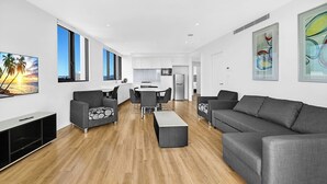Un salon moderne avec un canapé, un fauteuil et un téléviseur, un coin repas avec une table et des chaises, et une cuisine en arrière-plan.