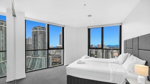 Une chambre d’hôtel moderne avec un grand lit, deux fenêtres donnant sur la ville, une table de chevet avec une lampe et un lit parfaitement fait.
