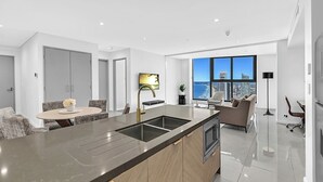 A modern kitchen with a large island, a dining area with a round table, and a living space with a sofa and a large window offering a city view. - Ocean and City View | 3BR Beachside Retreat Level 63 (Surfers Paradise)