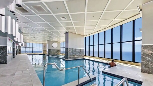 An indoor swimming pool with clear views of the ocean through large windows, a tiled floor, and a modern ceiling design. - Ocean and City View | 3BR Beachside Retreat Level 63 (Surfers Paradise)