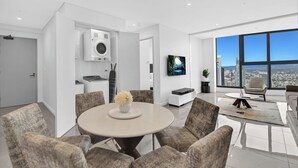 A modern living room with a round table, grey upholstered chairs, a flat-screen TV, and a city view through large windows. - Ocean and City View | 3BR Beachside Retreat Level 63 (Surfers Paradise)
