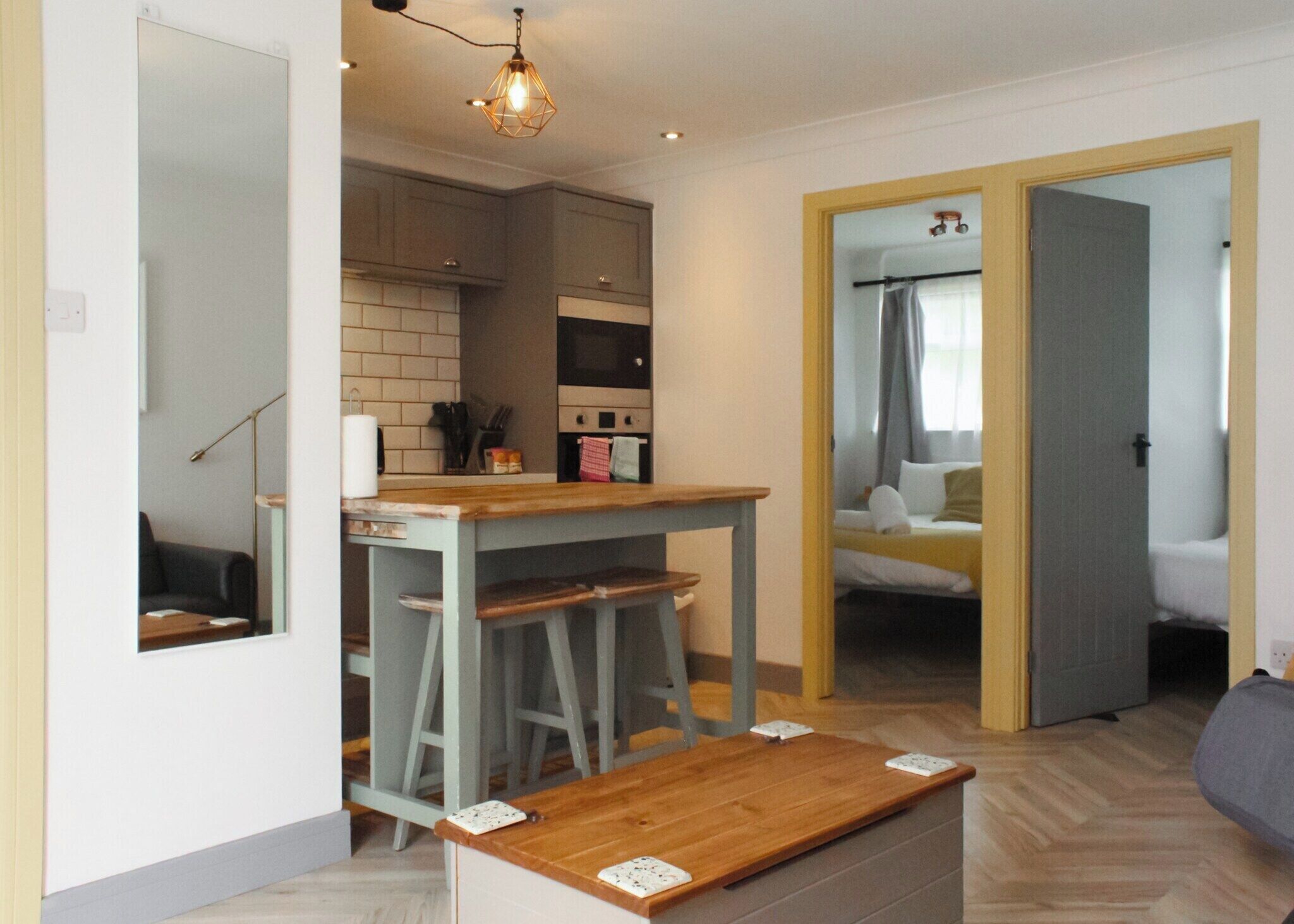 A compact kitchen with a wooden table and stools, a hanging light fixture, and a view into a bedroom through an open door.