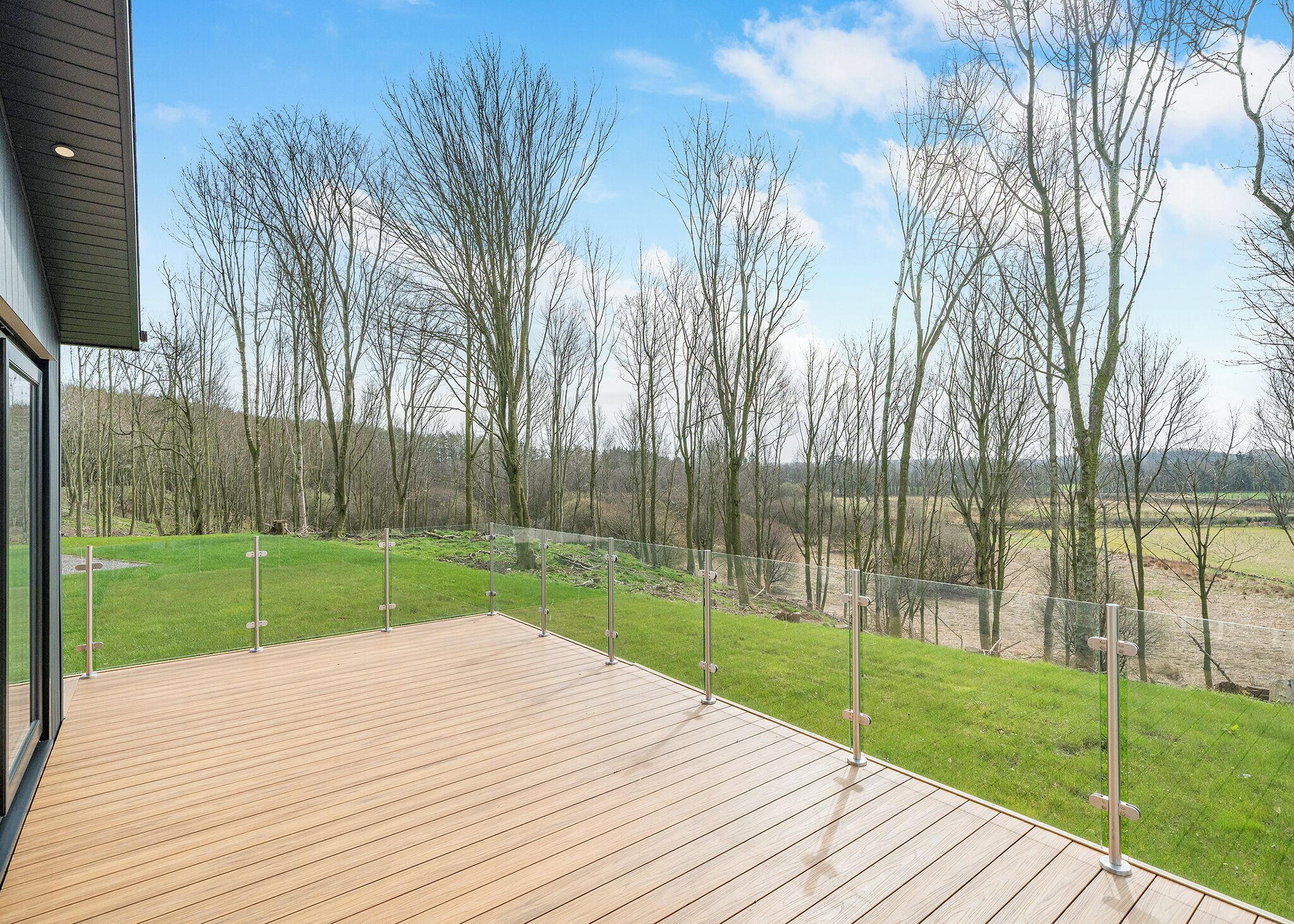 A wooden deck with a glass railing overlooking a grassy area with trees.