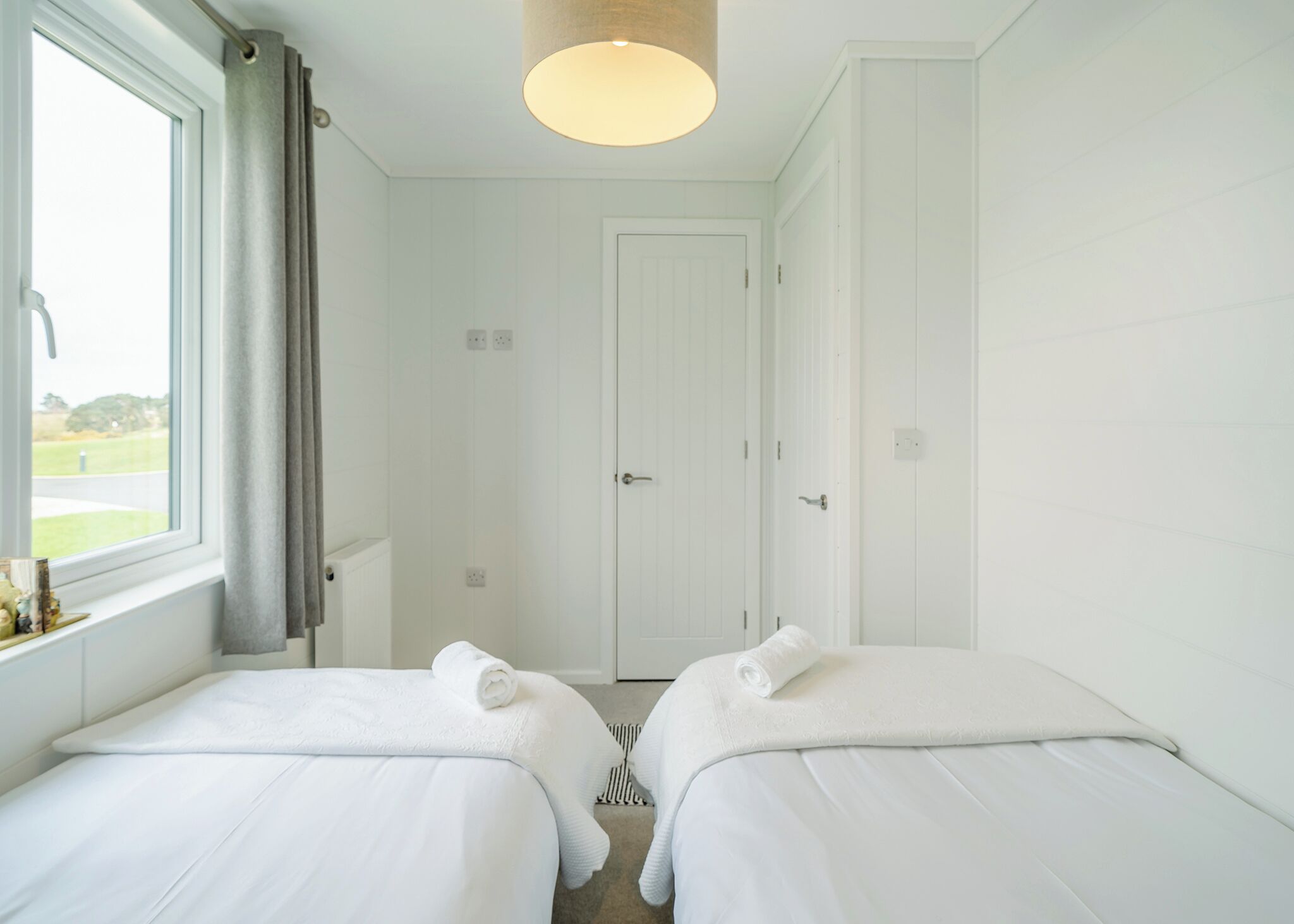 A hotel room with two beds, a window with curtains, a door, and a hanging light fixture.