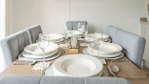 Una mesa de comedor dispuesta con platos, vasos y cubiertos, rodeada de sillas tapizadas de color gris, con un cuadro en la pared superior.