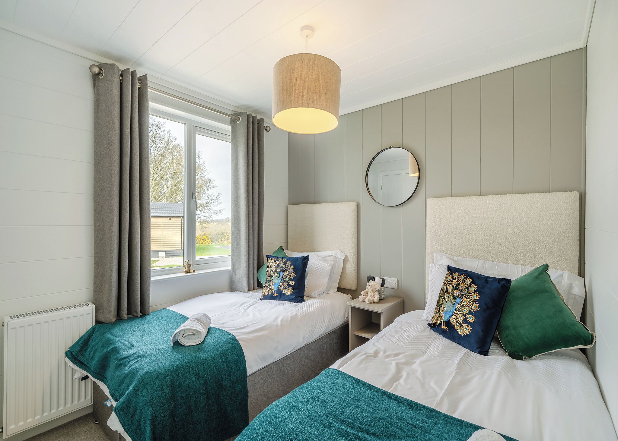 A bedroom with two beds, a window with curtains, a bedside table with a lamp, and a mirror on the wall.
