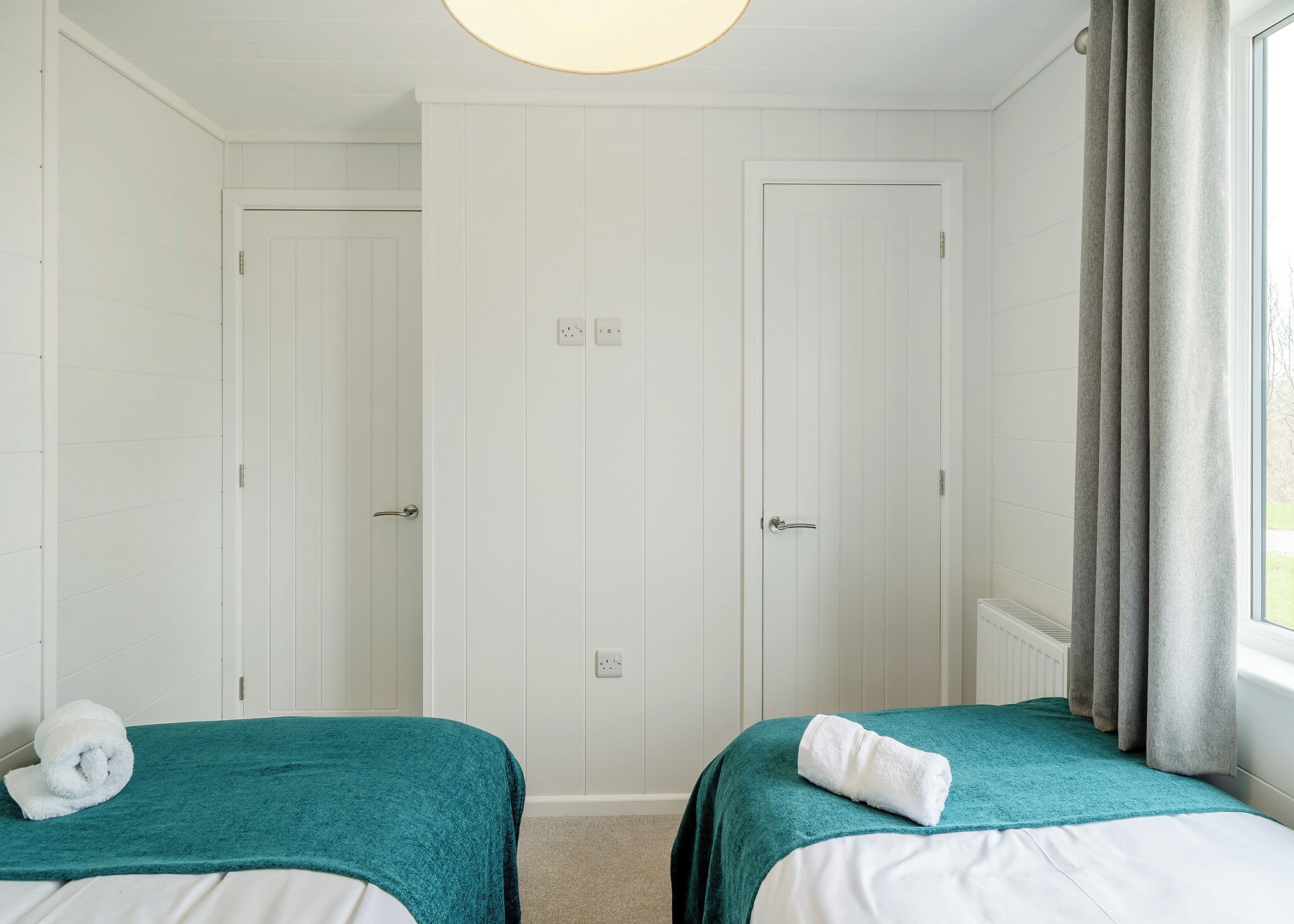 A hotel room with two beds, white walls, a window with curtains, and a ceiling light.