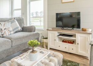 Uma sala de estar com um sofá cinzento, uma pequena planta em vaso, um rack branco com uma televisão e uma mesa de centro de madeira.
