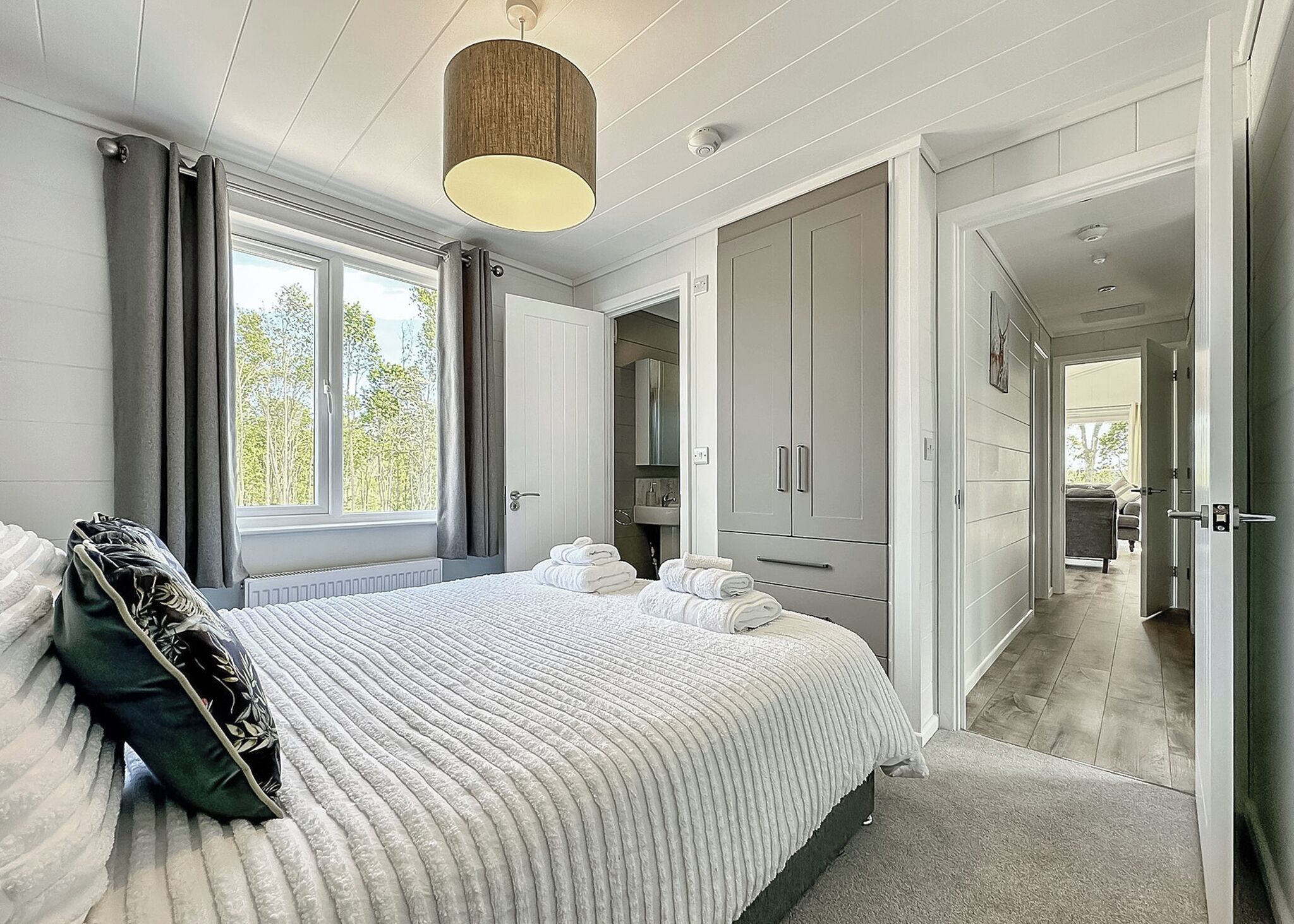 A bedroom with a bed, a bedside table, a window with curtains, a wardrobe, and a bathroom visible in the background.