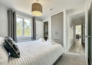 A bedroom with a bed, a bedside table, a window with curtains, a wardrobe, and a bathroom visible in the background.