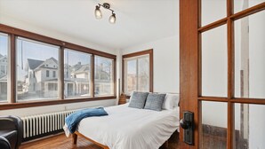A bedroom with a large bed, wooden floor, and a view of houses through the windows. - Urban Roots — Mins from Cedar Point.  (Sandusky)