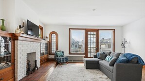 A living room with a fireplace, a sofa, a chair, and large windows. - Urban Roots — Mins from Cedar Point.  (Sandusky)