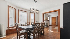 A dining area with a wooden table and chairs, large windows with blinds, and a hanging light fixture. - Urban Roots — Mins from Cedar Point.  (Sandusky)