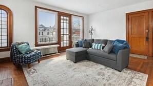 A living room with a grey sectional sofa, a patterned armchair, and a view of a house through large windows. - Urban Roots — Mins from Cedar Point.  (Sandusky)