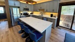 A modern kitchen with a large island, grey cabinets, stainless steel appliances, and blue bar stools. - Luxury Muskoka River Log Cabin (Bracebridge)