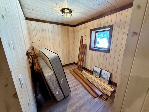 Habitación con paredes de madera, techo inclinado y una ventana. Hay colchones y tablones de madera en el suelo.
