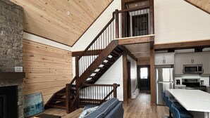 A modern loft-style interior with a wooden staircase, a kitchen area, and a seating area with a sofa and bar stools. - Luxury Muskoka River Log Cabin (Bracebridge)