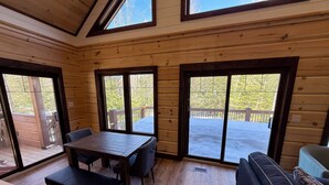 A wooden cabin interior with a dining table, chairs, a sofa, and large windows offering views of a deck and trees. - Luxury Muskoka River Log Cabin (Bracebridge)