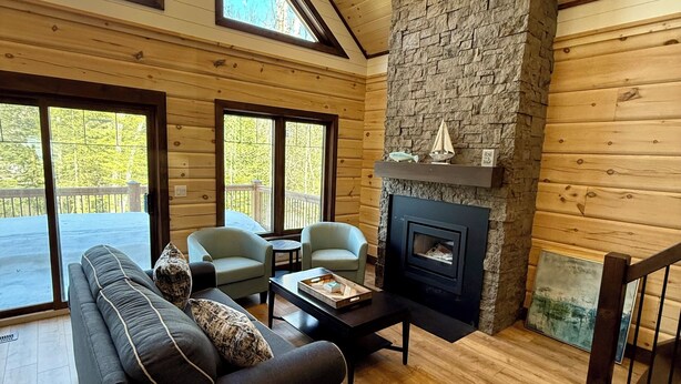 A spacious living room with a stone fireplace, wooden walls, and large windows. - Luxury Muskoka River Log Cabin (Bracebridge)