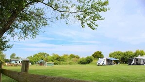 Ein grünes Feld im Vordergrund, mit einem Holzzaun, ein kleines Gebäude und mehrere Wohnmobile im Hintergrund.