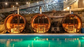 Trois pièces en forme de tonneau, construites en bois et dotées de murs en verre, une piscine et un mur de pierre en arrière-plan.