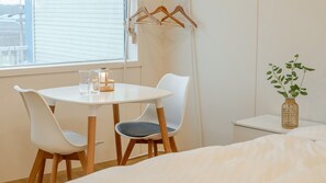 Een kamer met een ronde tafel, twee stoelen, een kledingrek, een klein nachtkastje met een plant en een raam met jaloezieën.