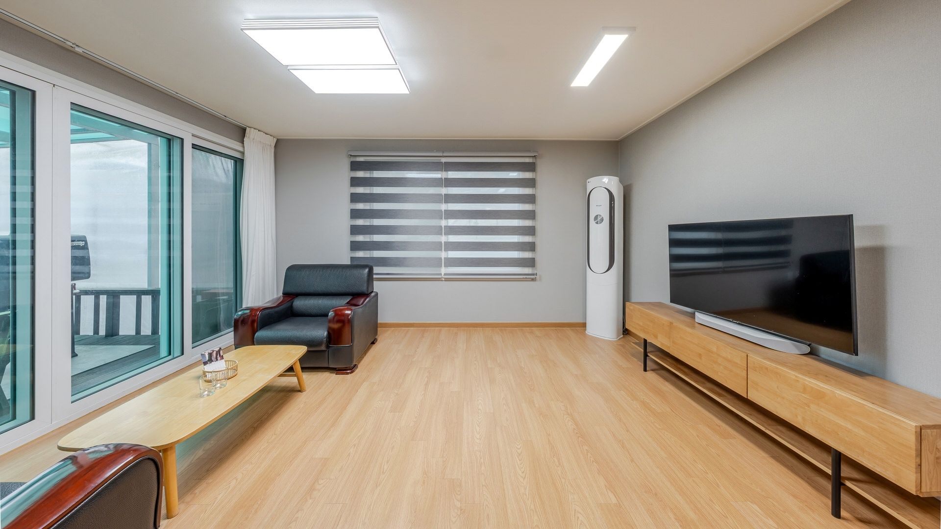 A modern living room with a large flat-screen TV, a leather armchair, a wooden coffee table, and a large window with blinds.