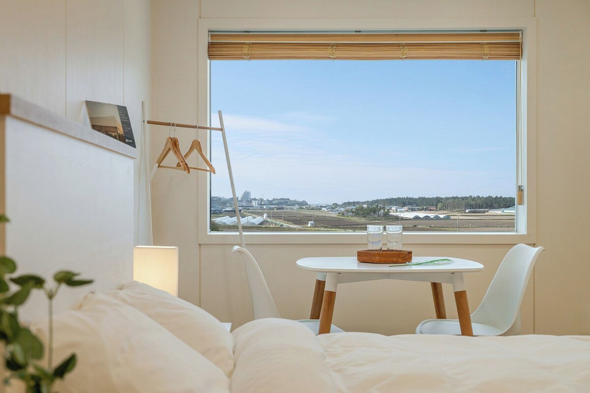 Une chambre d’hôtel avec un lit, une table et des chaises, une fenêtre donnant sur un aéroport et un luminaire suspendu.